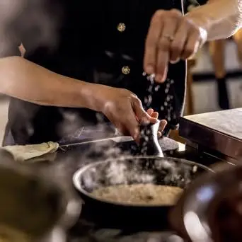 Hände streuen Salz über eine dampfende Pfanne mit kochendem Gericht; Dampf steigt auf.
