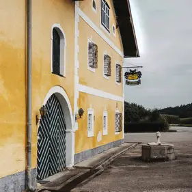 Gelbes Haus mit gemustertem Tor, Schild "Gasthaus" und kleinem Steinbrunnen an der ruhigen Straße.