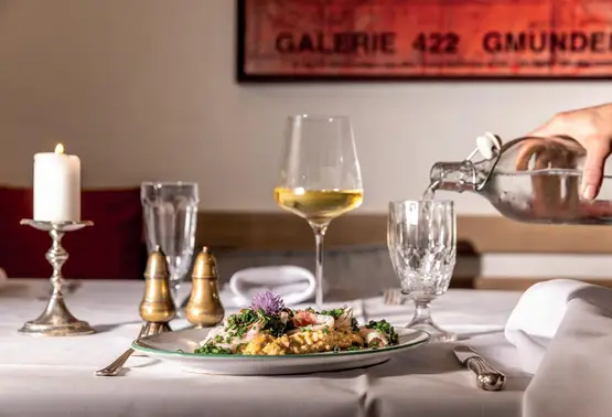 Risottoteller auf weißem Tischtuch, Weinglas und Kerze; Hand gießt Wasser, im Hintergrund GALERIE 422 GMUNDEN.